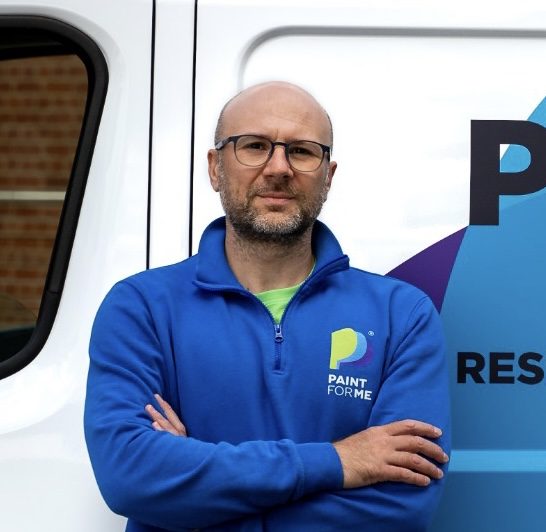 The business owner in a blue sweatshirt stands confidently with crossed arms beside a vehicle.